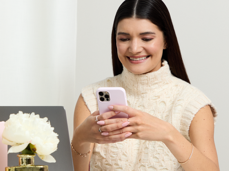 Person seated at a desk using a smartphone beside a laptop and flowers, wearing a light textured top in a bright, neutral workspace.